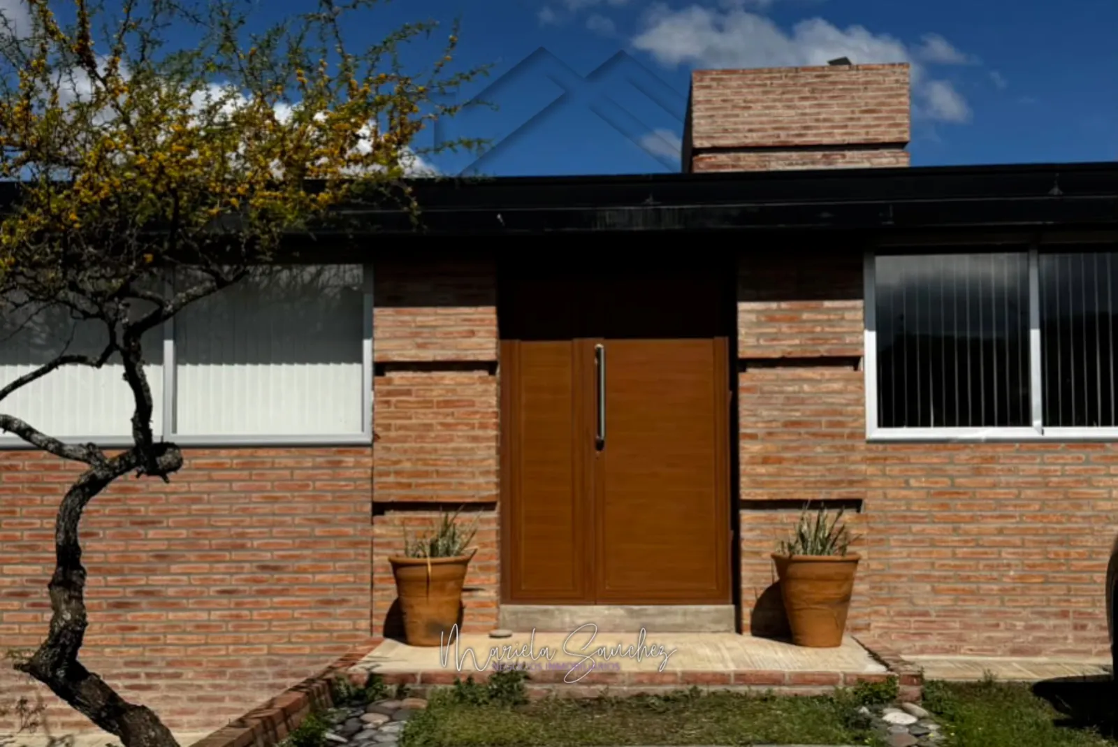 Frente de ladrillo visto de la casa, con la puerta de madera entre dos macetas de barro